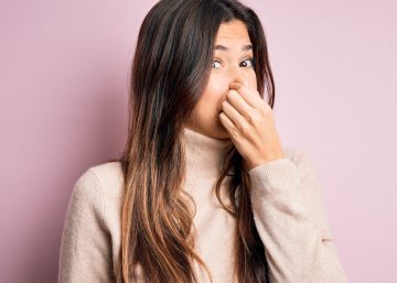Young woman wearing sweater standing in front of pink background Holding Breath With Fingers On Nose.