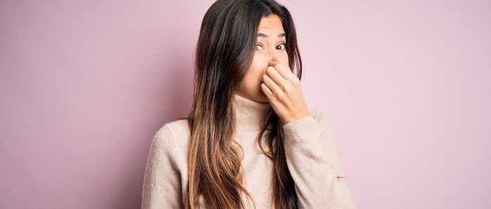 Young woman wearing sweater standing in front of pink background Holding Breath With Fingers On Nose.