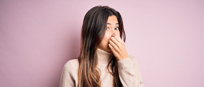 Young woman wearing sweater standing in front of pink background Holding Breath With Fingers On Nose.
